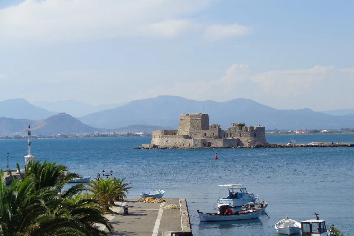 A small stone fort with a round tower occupying the whole of a tiny islet in a calm harbour, with hills on the far shore behind it.