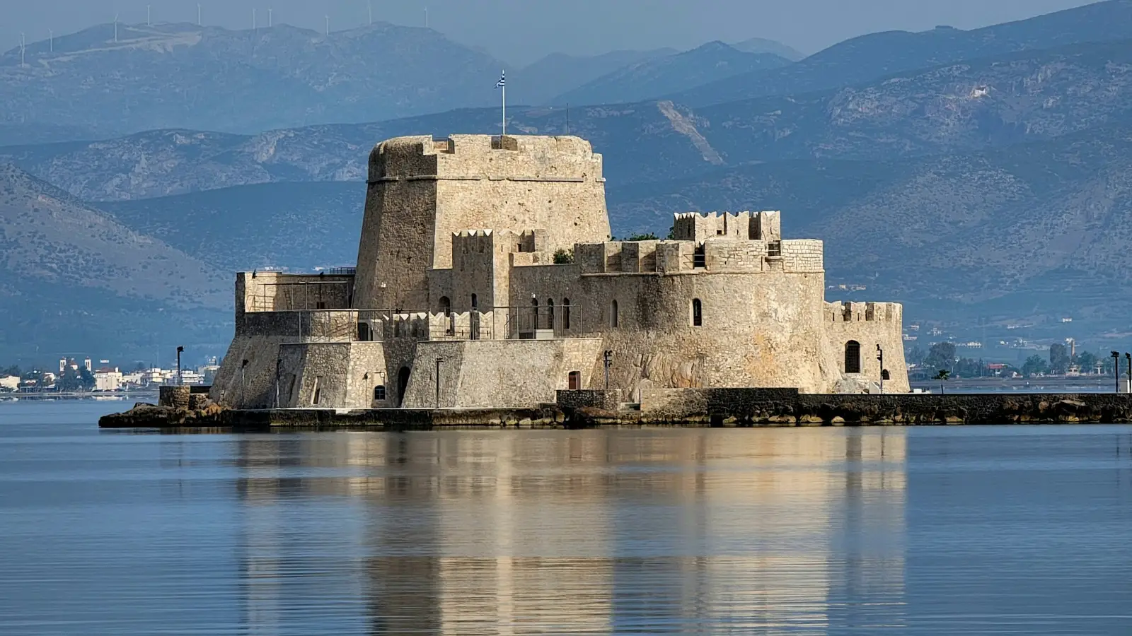 The Bourtzi of Nafplio at first light: the fortified islet standing alone in the harbour, its tall polygonal keep rising above crenellated curtain walls and rounded bastions, the water completely surrounding it and mirroring it, and the hills of the Argolid behind