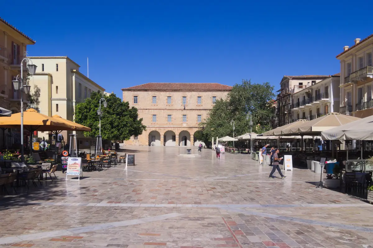 A wide paved square in an old town, ringed by two- and three-storey stone and neoclassical buildings, with a large arched stone building closing the far end.