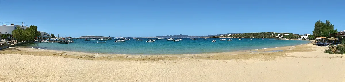 Alyki bay on the south coast of Paros: an empty sweep of pale sand, flat turquoise water with small boats at their moorings, and low hills closing the bay