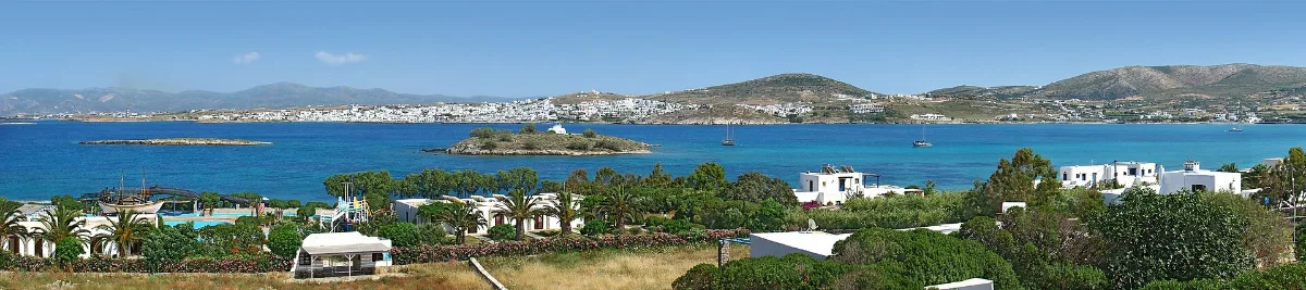 Naoussa on Paros seen across its bay: a low white town on the far shore, brown hills behind, a small islet with a white chapel in the middle of turquoise and blue water