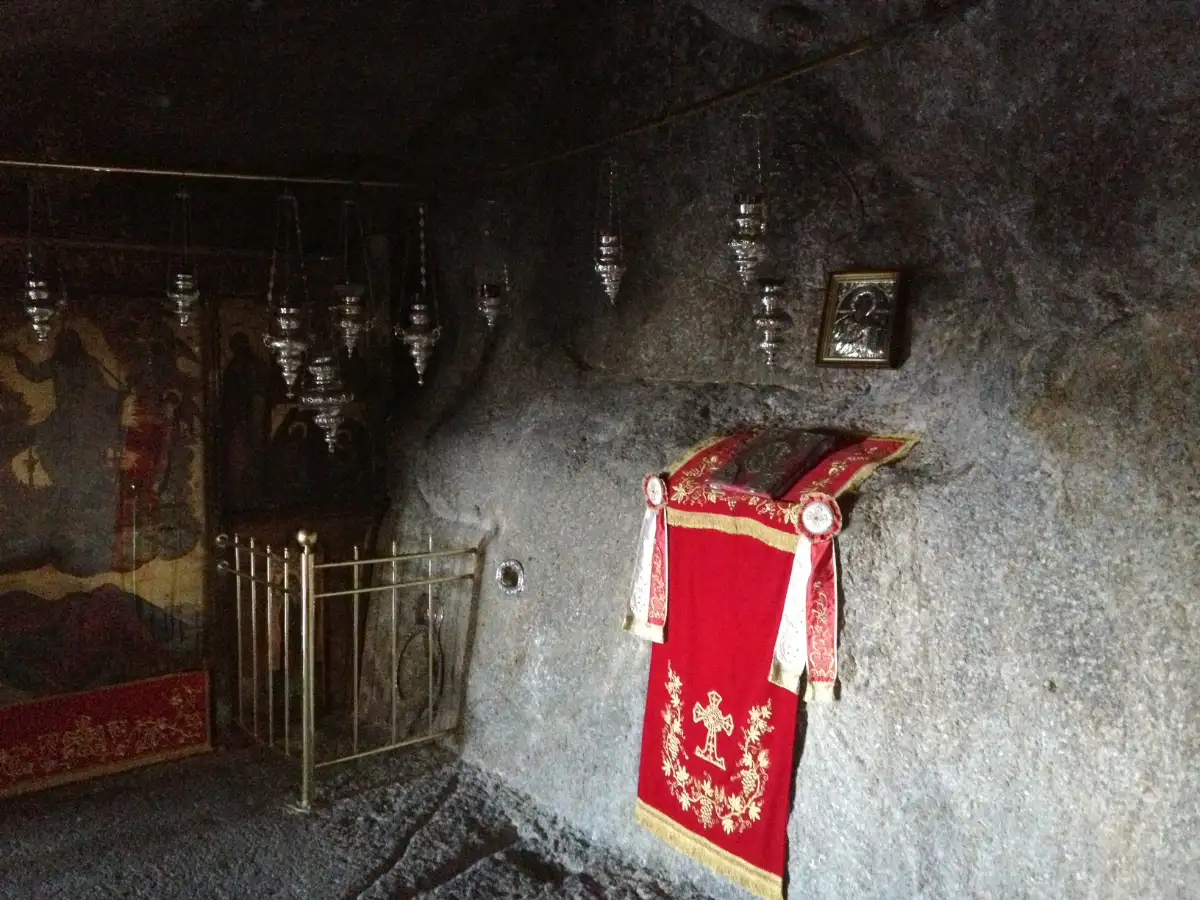 The inside of a small rock grotto fitted out as a chapel: bare grey rock walls and ceiling, a row of hanging silver oil lamps, painted icons set against the rock, and a brass rail around a hollow in the rock floor.