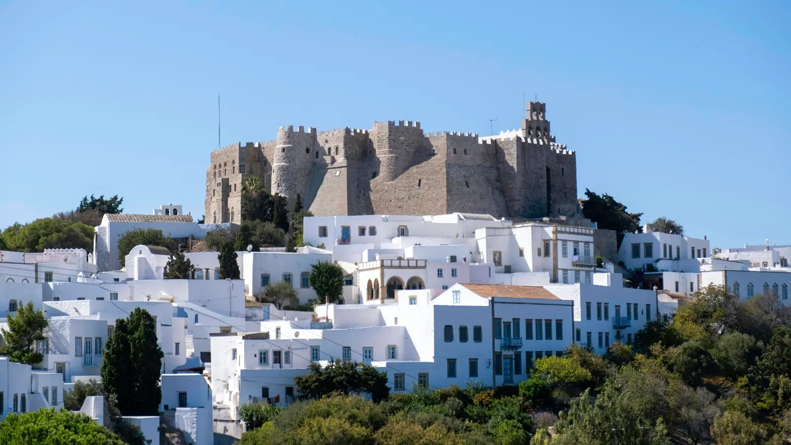 The fortress-monastery of Saint John the Theologian standing on the hilltop above Chora on Patmos: the dark battlemented monastery walls crowning the ridge, the whitewashed houses of Chora packed on the slope below it