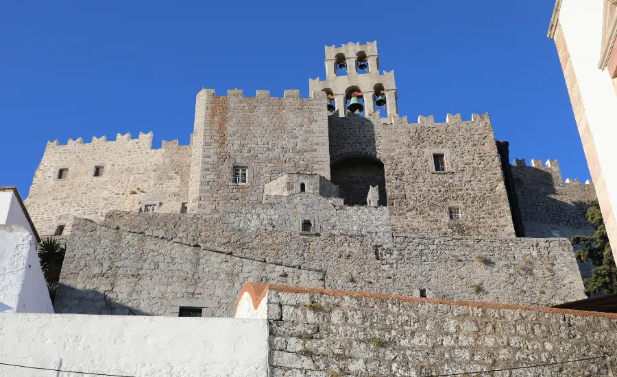 A tall fortified stone building rising above whitewashed houses, its walls topped with square crenellations and pierced by small deep-set windows, with a triple bell gable carrying three bells above the roofline.