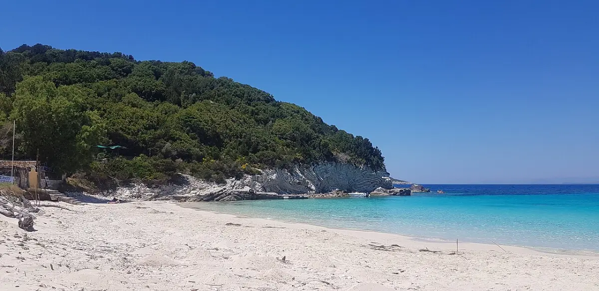 Vrika beach on Antipaxos: empty pale sand and clear turquoise water beneath a green pine-covered headland with white rock, under a deep blue sky