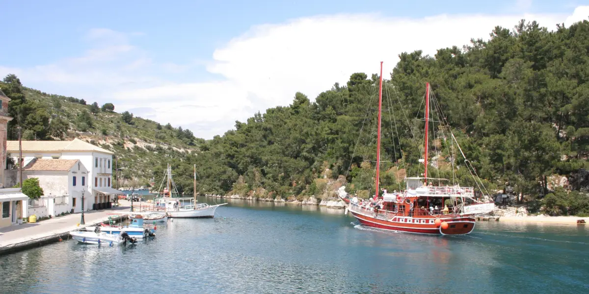 A narrow channel of flat green water between a low quay with pastel buildings on one side and a steep pine-covered islet on the other, with small fishing boats moored and a wooden excursion boat under way.