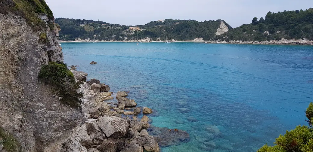 A sheltered bay of pale turquoise water shading to deep blue, ringed by low green hills and white rock, with a handful of yachts anchored off a small beach at the head of it.