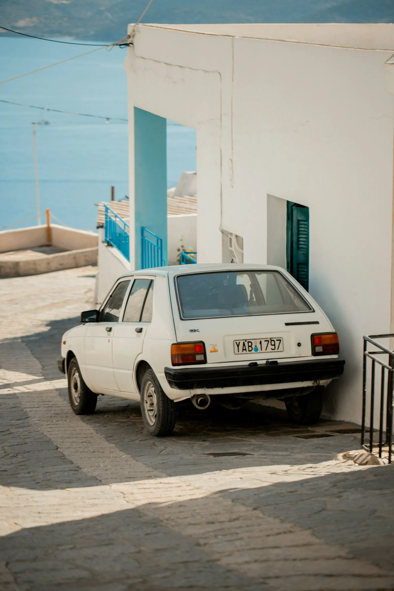 A small white car parked on a village street on Milos, with the sea beyond