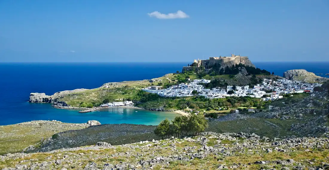 Lindos on Rhodes seen from a ridge: the walled acropolis on its rock, the white village below it and a turquoise bay at the foot of the slope