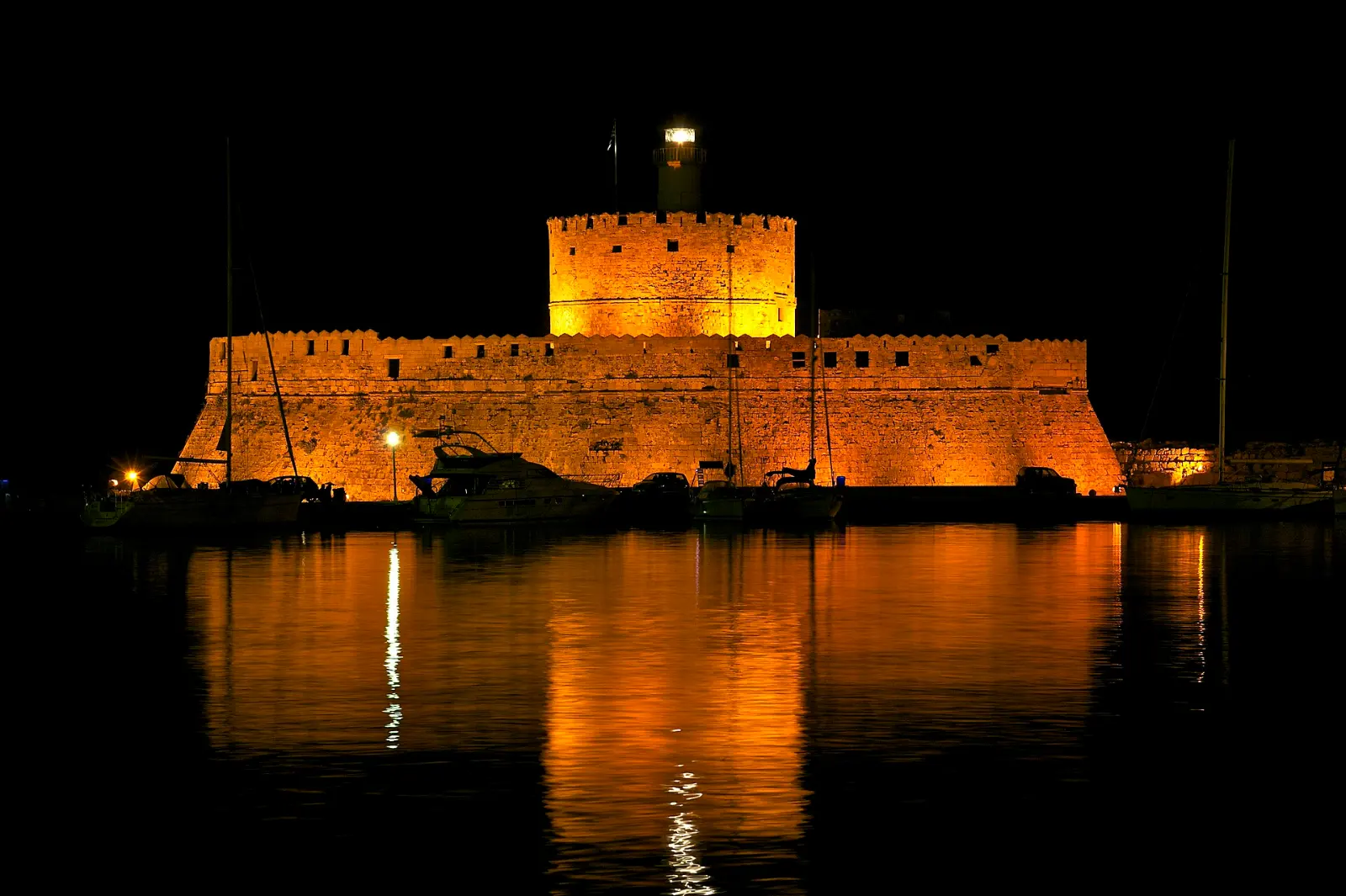 The floodlit Fortress of Agios Nikolaos and its lighthouse at Mandraki harbour, Rhodes, reflected in still black water at night