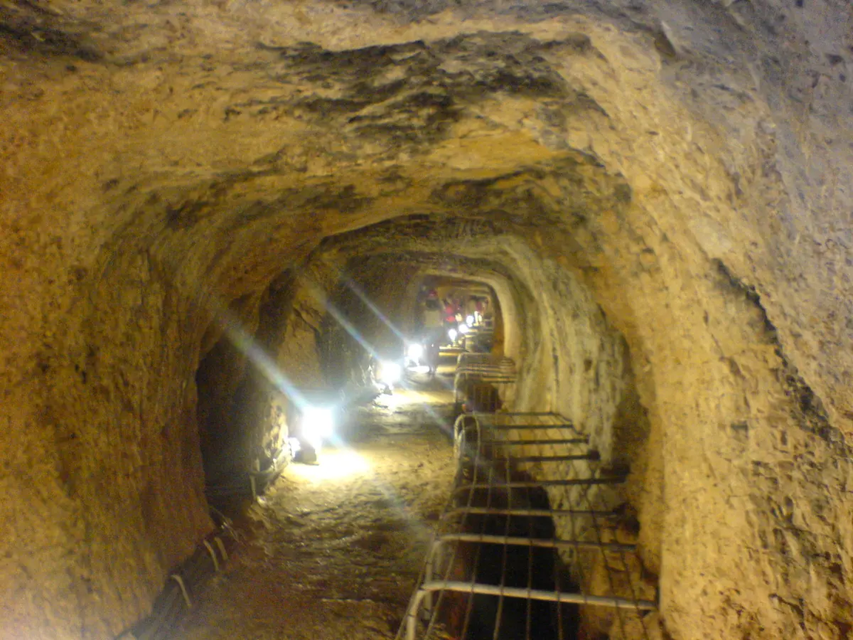 A rough rock-cut tunnel running straight into the distance, its roof roughly arched, lit by lamps set along the floor, with a metal handrail on the right and a narrow channel beside the walkway.