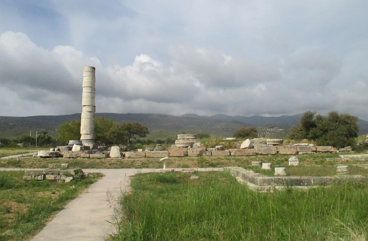 A flat archaeological site under a cloudy sky: one tall column of stacked marble drums standing alone, with low foundation courses and scattered stone blocks laid out across the grass around it and hills behind.