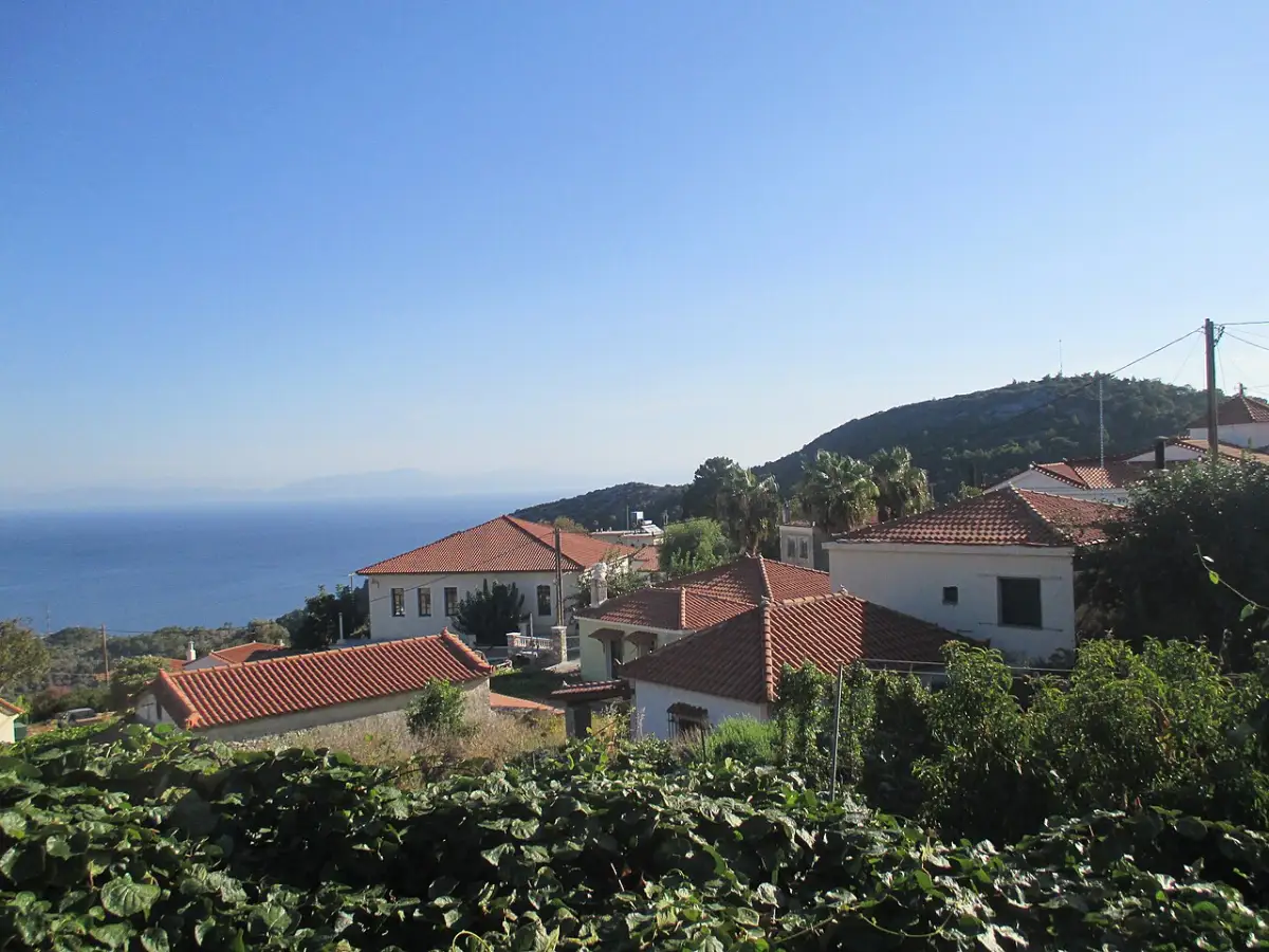 The mountain village of Vourliotes on Samos: white houses with red-tiled roofs on a green forested slope above a blue sea, under a clear sky
