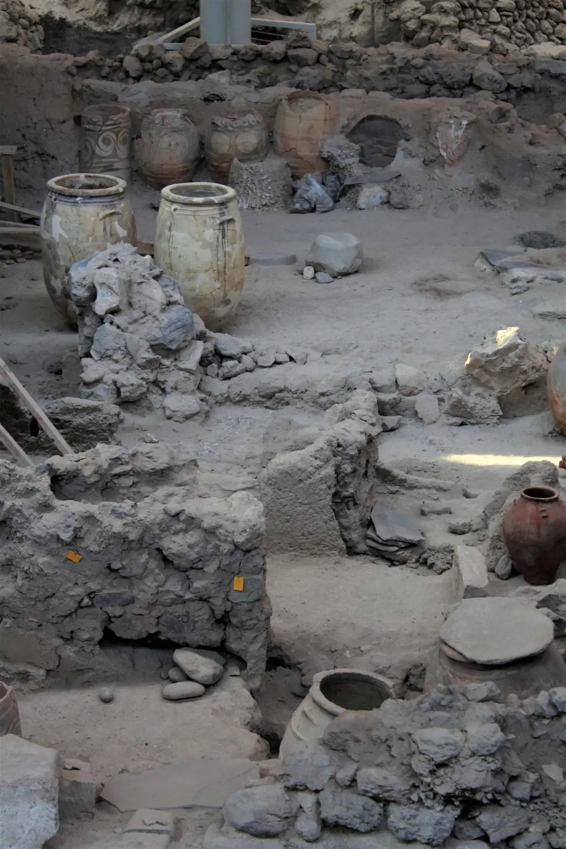 Excavated Bronze Age walls and large Minoan storage jars standing among them at the Akrotiri site on Santorini, under the modern roof that shelters the dig