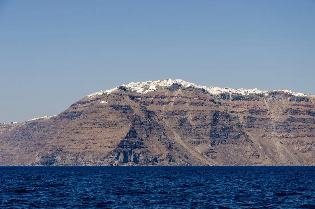 The Santorini caldera cliff seen from the water, layered volcanic rock with a line of white village buildings along the rim
