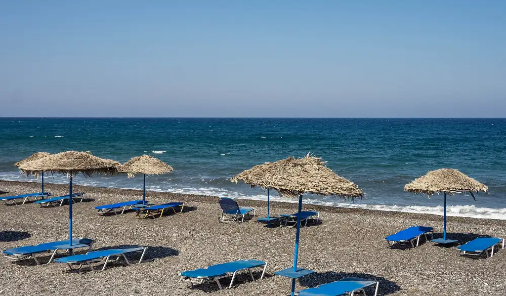 Empty blue sunbeds and straw umbrellas on the dark volcanic grit of Kamari beach, Santorini, with the sea beyond