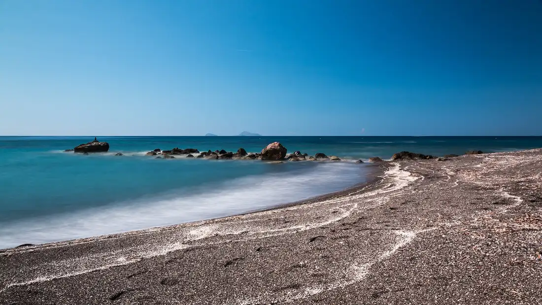 The empty dark gravel shore at Vlychada on Santorini’s south coast, with boulders in shallow turquoise water under a deep blue sky