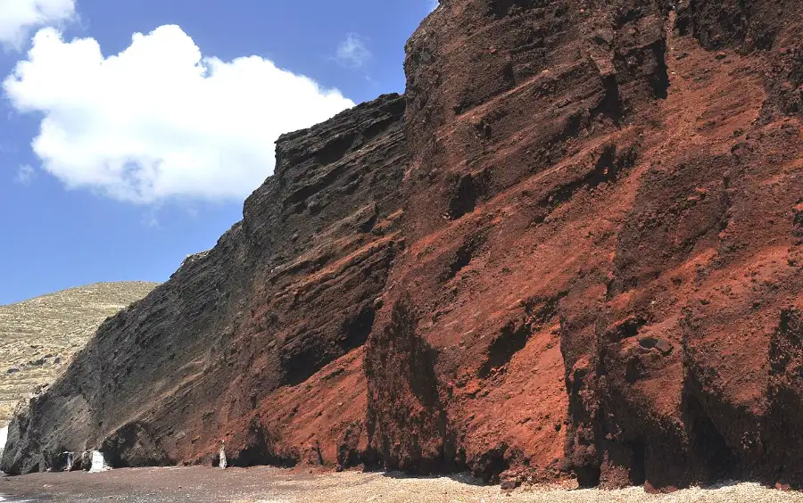 A Santorini shoreline of dark coarse volcanic pebble beneath a red-brown cliff