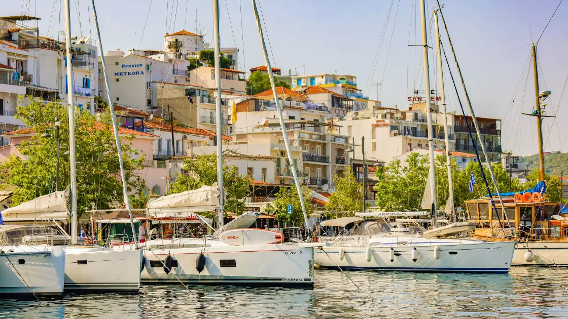 The old harbour of Skiathos Town: a row of moored yachts and sailing boats with bare masts in the foreground, and behind them the waterfront rising in tiers of white and pastel houses with terracotta roofs, balconies and quayside tavernas among plane trees