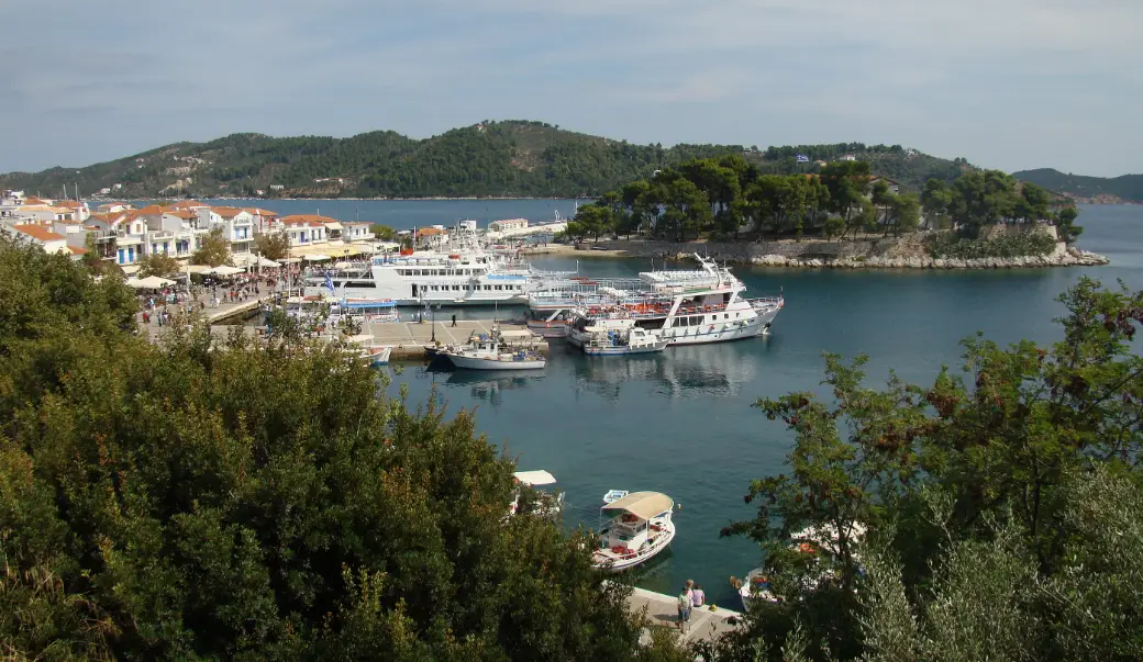 A harbour seen from above: white houses with red-tiled roofs and a waterfront of awnings on the left, passenger ferries and fishing boats moored at the quay, and a small pine-covered headland projecting into the water in the centre.