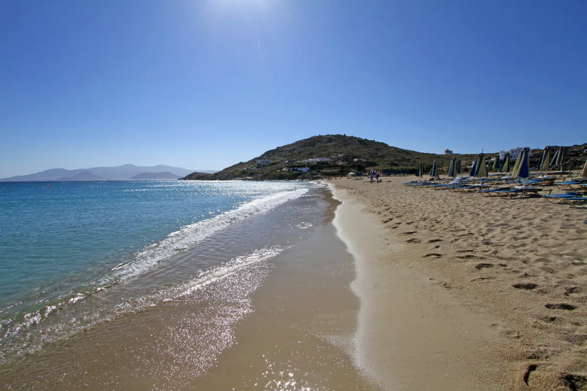 A wide sandy beach on Naxos with rows of closed umbrellas and sun loungers set back on the dry sand at one side, and open free public sand with a clear empty waterline at the other