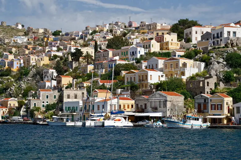 Tall houses in cream, ochre, pale yellow and blue, with pediments, shuttered windows and terracotta roofs, stacked in tiers up the steep rocky sides of a bay above a quay lined with fishing boats and yachts.