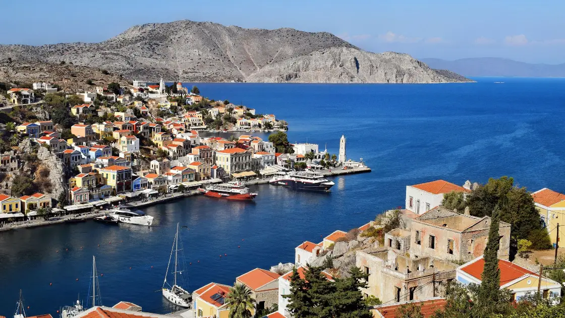 Gialos, the harbour of Symi, seen from the hillside above: tiers of neoclassical mansions in ochre, pink, cream and pale blue with pedimented facades and terracotta roofs stacked up both sides of a narrow inlet, the stone clock tower on the quay at the harbour mouth, and boats moored along the waterfront