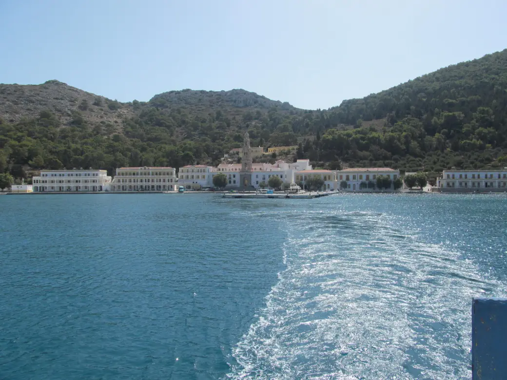 Long two-storey ranges of white and pale yellow buildings with rows of arched windows standing at the water's edge around the head of a sheltered bay, a tall bell tower rising above them, and wooded hills closing the bay on both sides.