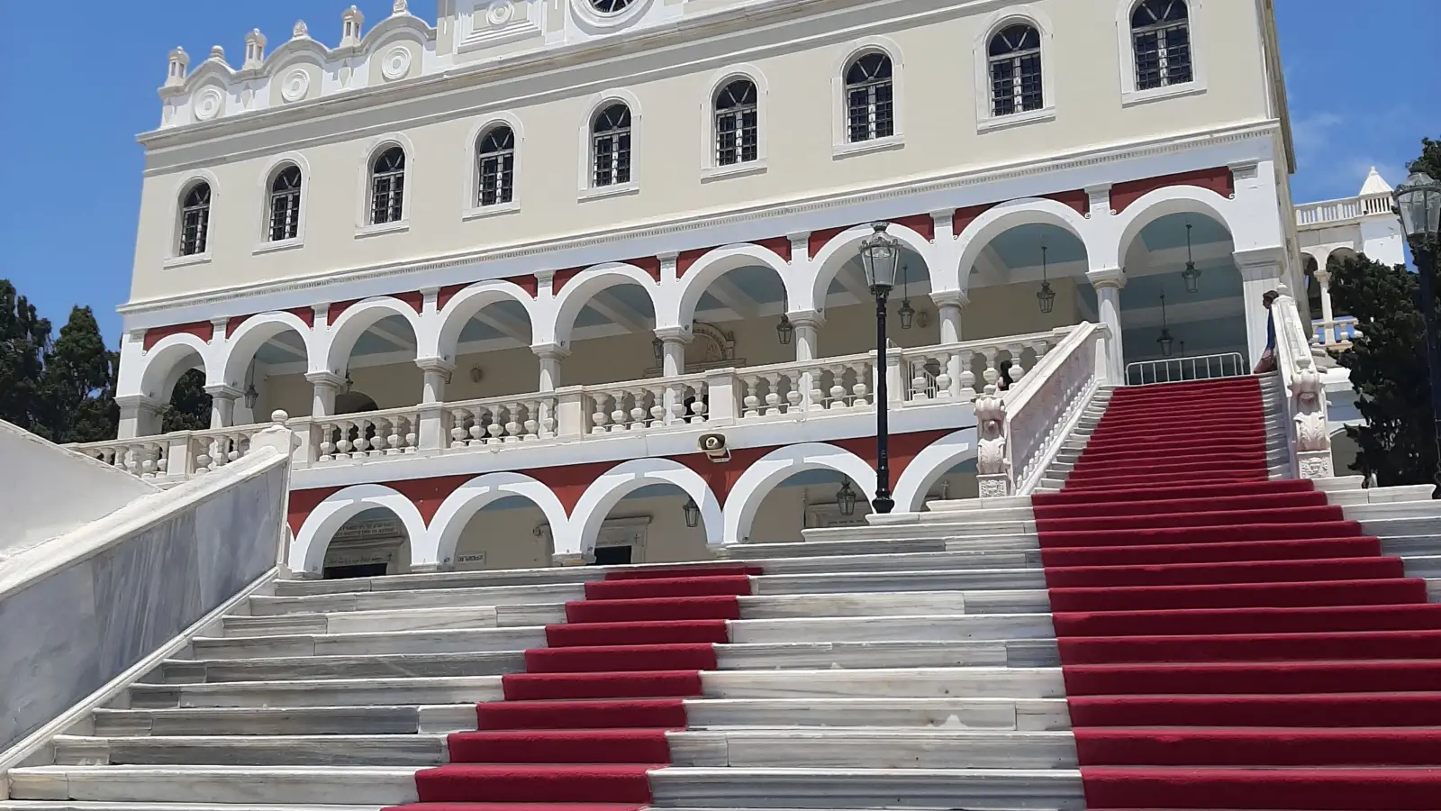 The church of Panagia Evangelistria in Tinos Town: the white marble facade with its arcaded front and bell tower, and the red carpeted steps running up the approach to the entrance, against a clear blue sky