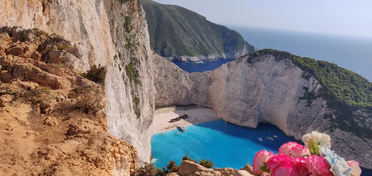 Navagio cove on Zakynthos seen from the clifftop - a rusted shipwreck on the sand of an enclosed beach ringed by tall white cliffs, with small boats on the turquoise water