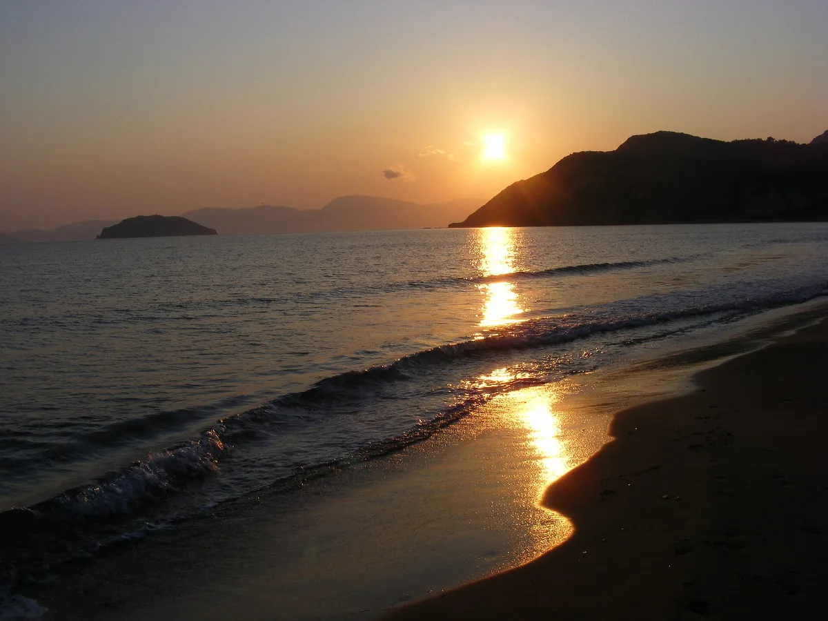 Gerakas beach on Zakynthos with the sun low over a dark headland, empty wet sand, small breaking waves and a gold light column on the water