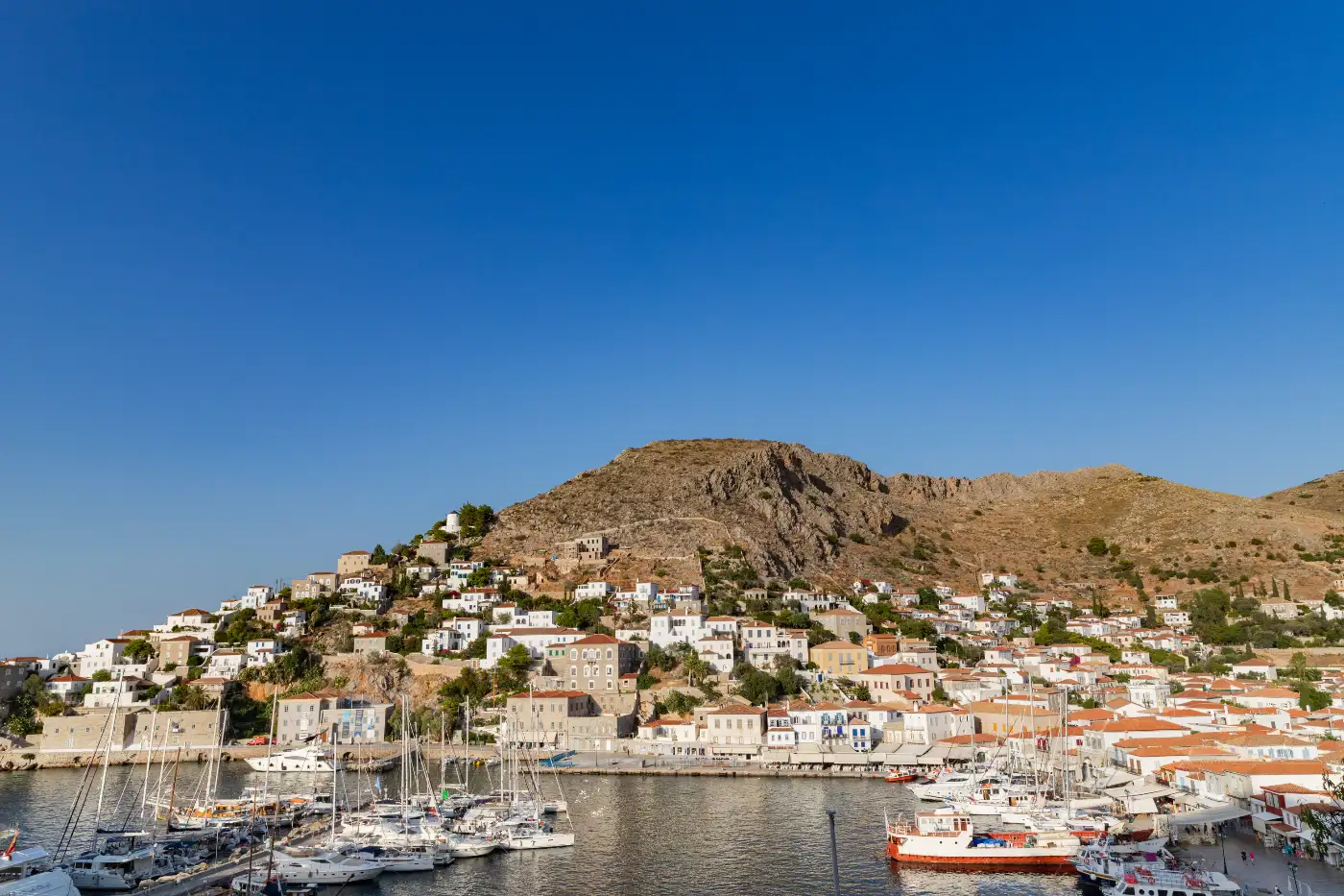 The harbour of Hydra, its stone houses rising in a horseshoe around the water, yachts and caiques moored along the quay and a bare rocky hill behind the town