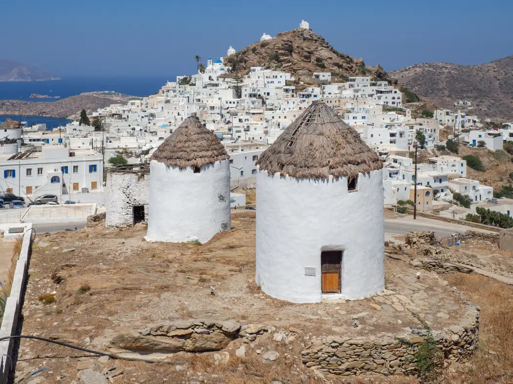 Two thatched whitewashed windmills on the ridge east of Chora on Ios, with the white village spread up its hill behind them, a blue-domed church partway up and a chapel on the rocky summit