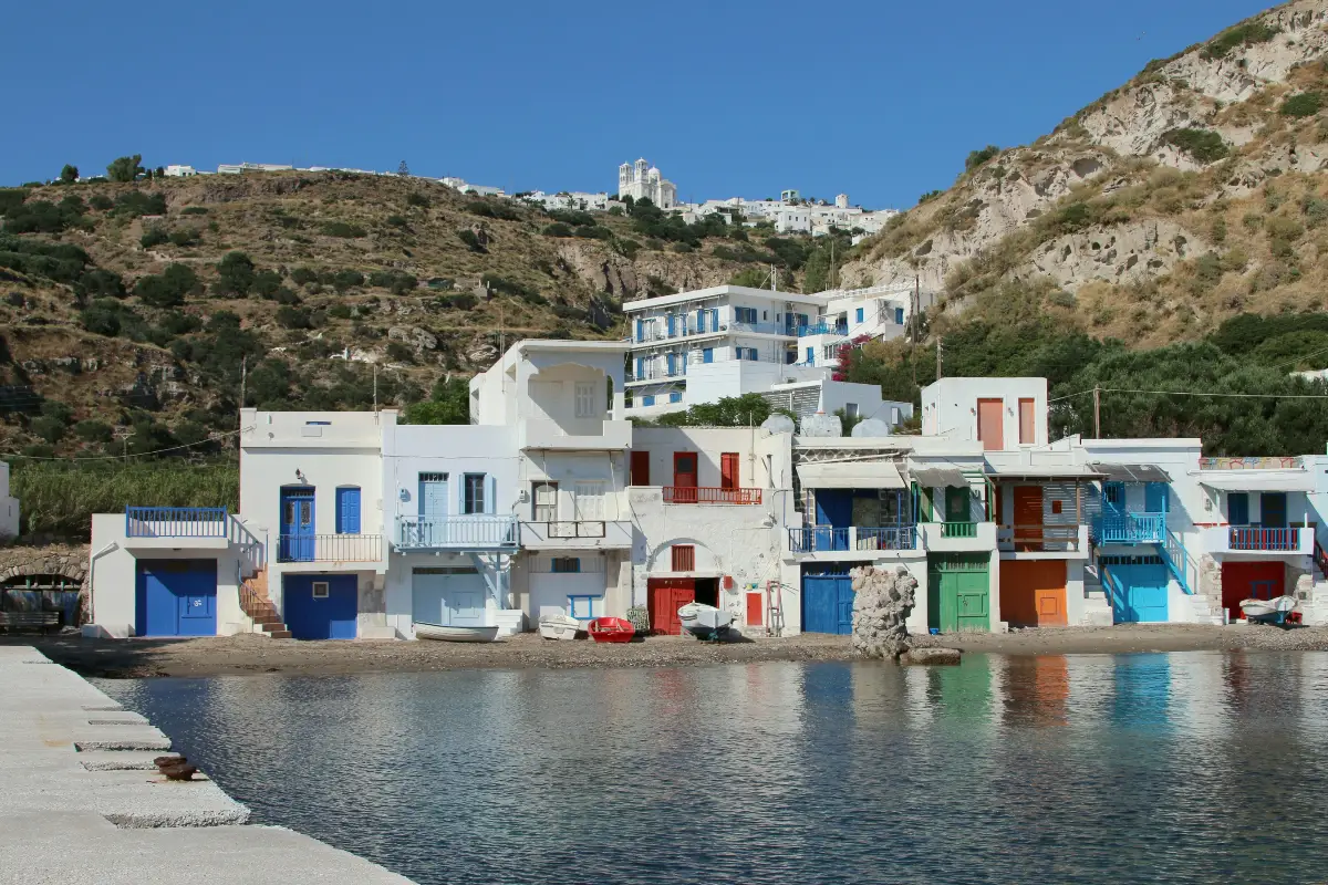 The row of two-storey syrmata boathouses at Klima on Milos, their garage doors and balconies painted in blue, red, green and orange, reflected in calm water, with Trypiti on the ridge above