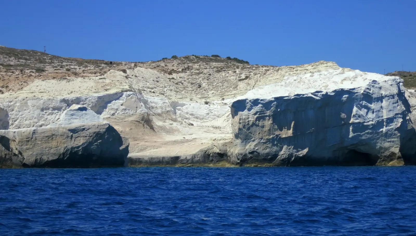 The white rock shelf of Sarakiniko on Milos seen from the water, its smooth pale formations undercut by a sea cave, with dark blue sea below and a low scrub ridge above