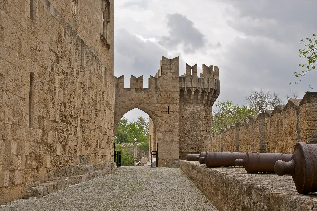 The Cannon Gate in the medieval walls of Rhodes Old Town, reached along an empty stone ramp beside the tall wall of the Grand Master's palace, with two old cannon barrels resting on the parapet