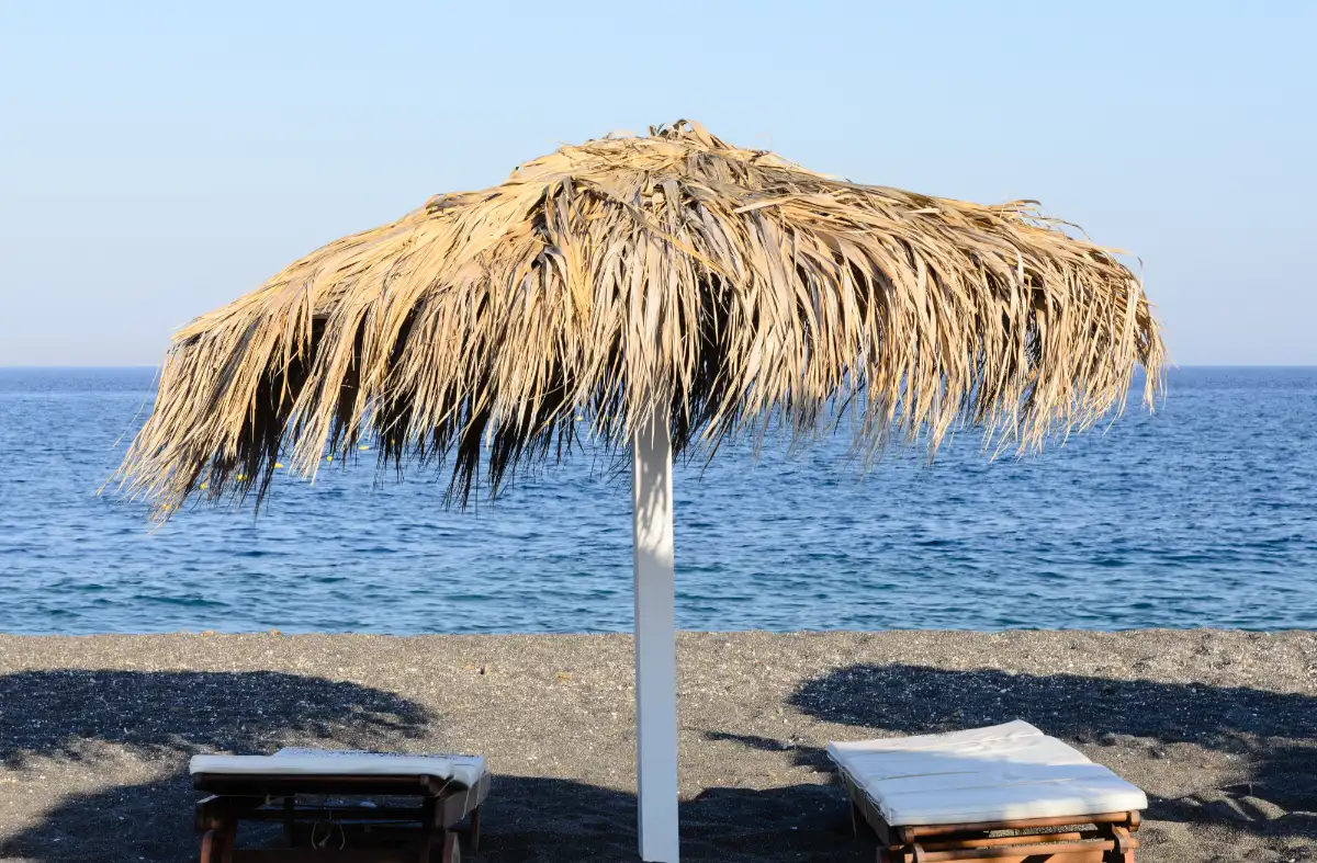 A thatched palm-frond parasol and two empty sunbeds on the black volcanic sand at Perissa on Santorini, with the sea beyond