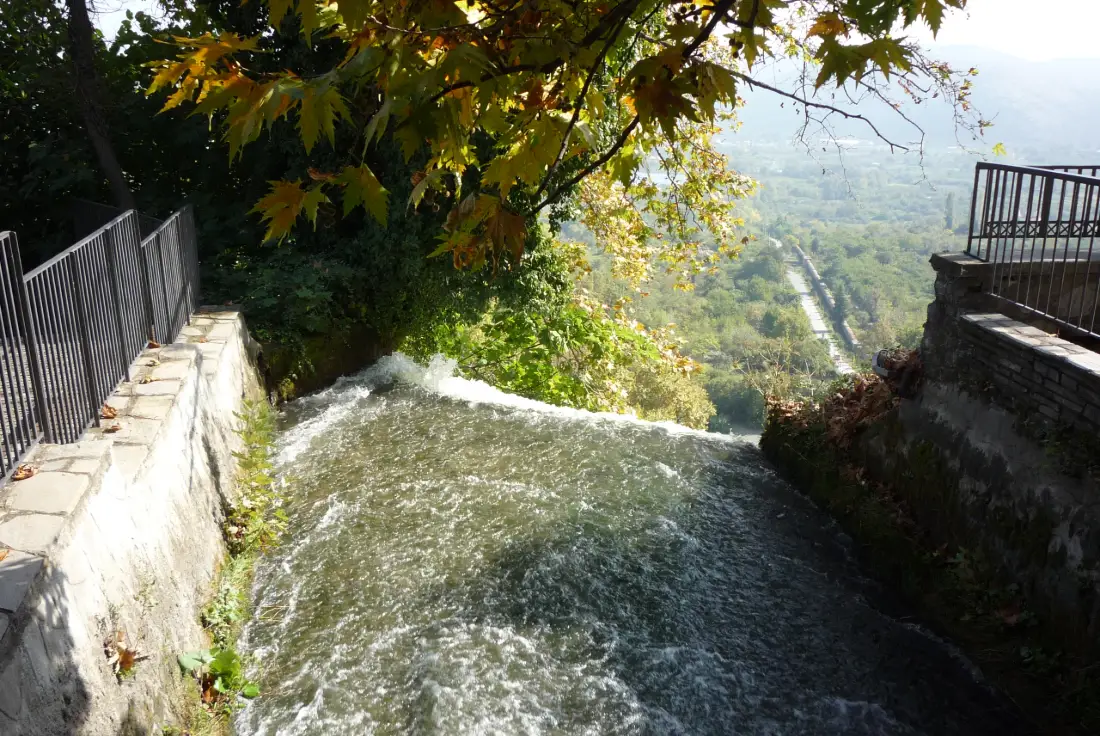 Water running fast down the channel above the Edessa falls, with railings alongside and the Macedonian plain beyond