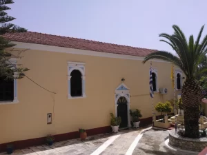 הכנסייה בספרטוכורי שבמגאניסי, מבנה בצהוב חיוור עם חלונות מקושתים ופתח לבן, דקל ודגלי יוון בכיכר המרוצפת שלפניה